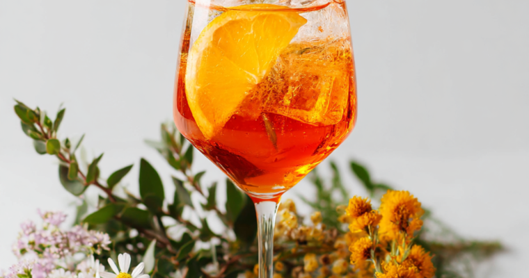 A refreshing glass of Aperol Spritz with ice, orange slices, and floral garnish on a white background